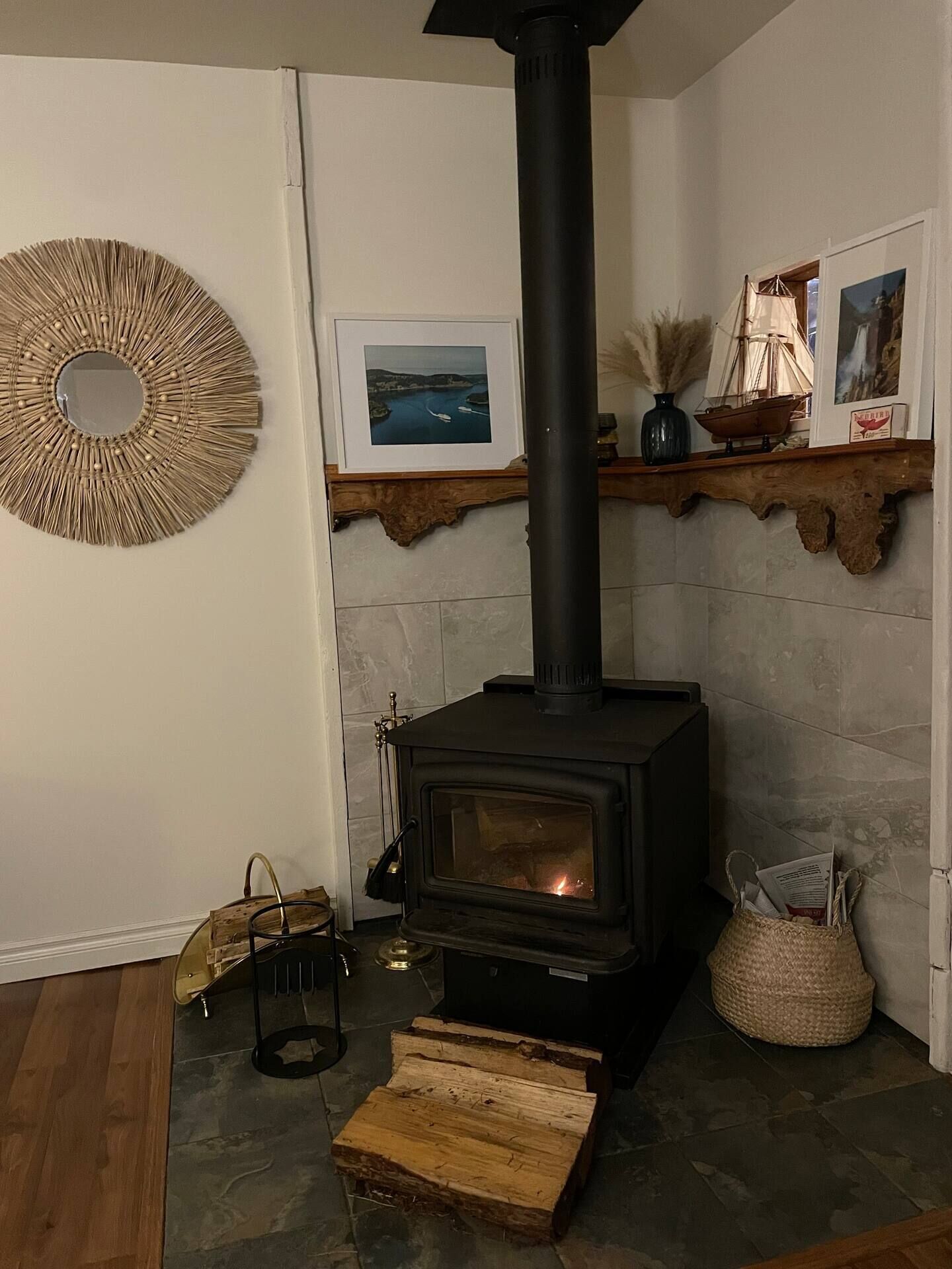 Een houtkachel met schoorsteen, een ingelijst landschapsbeeld en een decoratieve spiegel aan de muur.