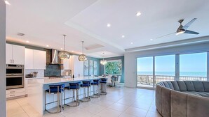 A modern kitchen with a central island, bar stools, and a large window overlooking the sea. - Beach One: Style Meets the Shore w/ Pool and Spa (Treasure Island)