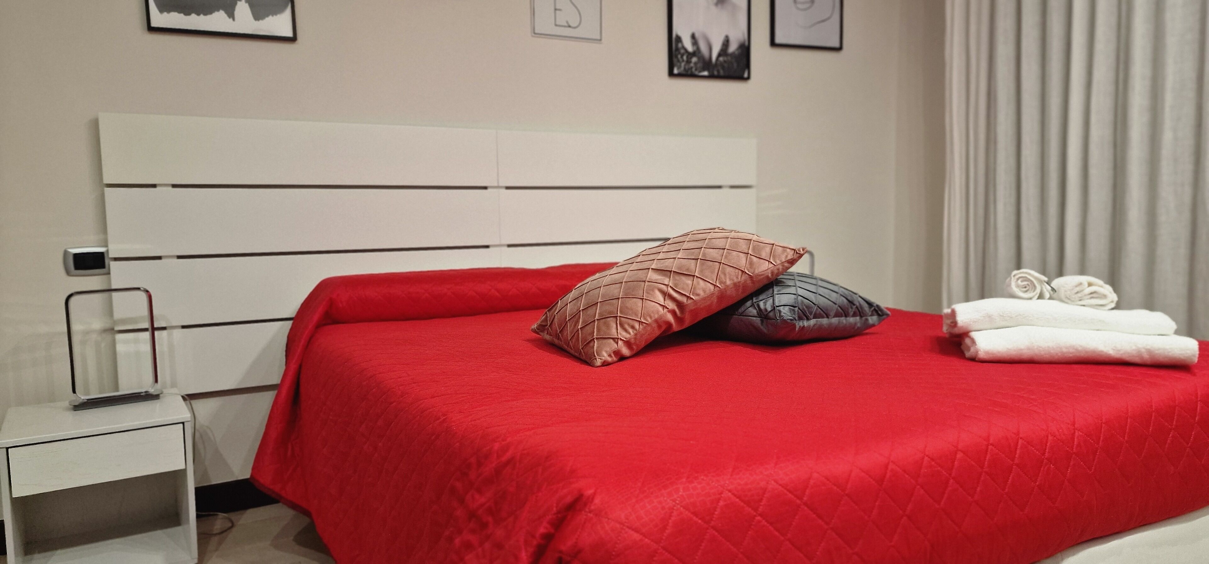 Un lit bien fait, recouvert d’une couverture rouge, agrémenté de deux coussins décoratifs et d’une pile de serviettes pliées posées sur une table de chevet blanche.