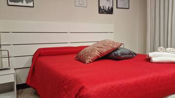 Un lit bien fait, recouvert d’une couverture rouge, agrémenté de deux coussins décoratifs et d’une pile de serviettes pliées posées sur une table de chevet blanche.