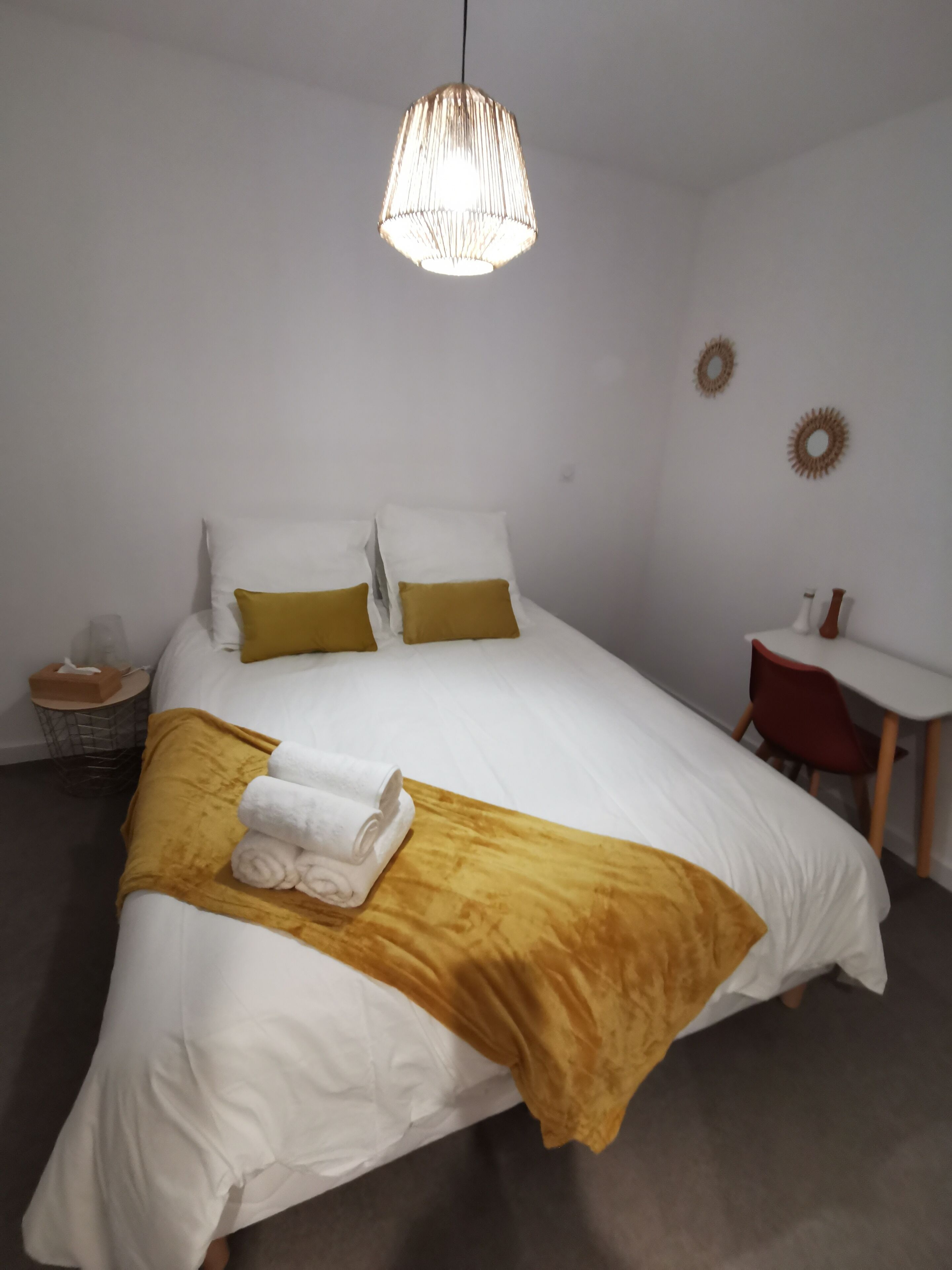 Una cama bien tendida con ropa de cama blanca y mostaza, una mesita de noche pequeña con un pañuelero y un escritorio con una silla.
