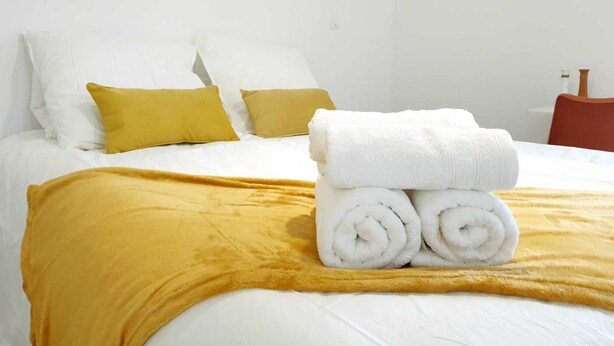 A neatly made bed with white linens, two mustard-colored pillows, and a rolled white towel on the bed. - The bohemian (Pamiers)