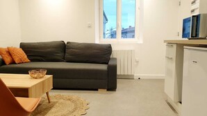 A compact living space with a dark grey sofa, a small wooden table, and a single orange cushion. A white radiator is visible under a window, and a white cabinet with a microwave is on the right. - The bohemian (Pamiers)