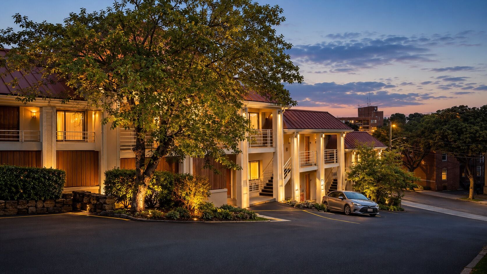 Un hotel de varios pisos con un patio central, un coche estacionado al frente y un árbol en primer plano.