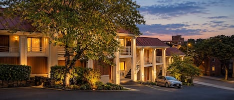 A multi-story hotel with a central courtyard, a car parked in front, and a tree in the foreground.