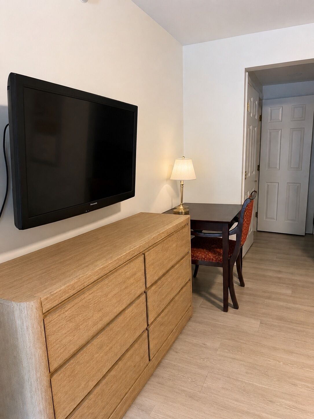 Een kamer met een wandgemonteerde flatscreen televisie, een houten dressoir met lades, een eettafel met stoelen en een lamp op het dressoir.