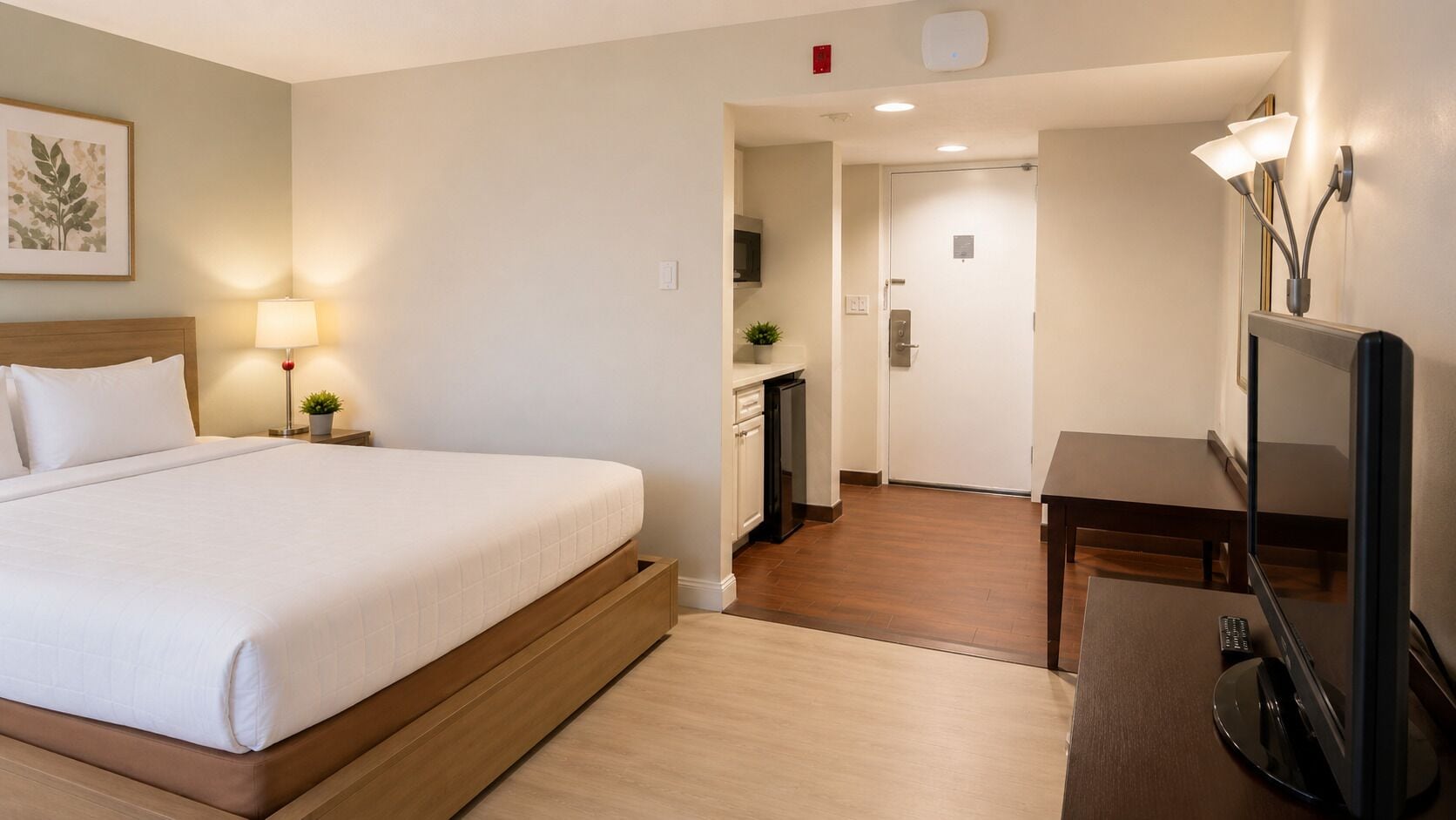 A hotel room with a bed, bedside table, lamp, TV, and a kitchenette area.