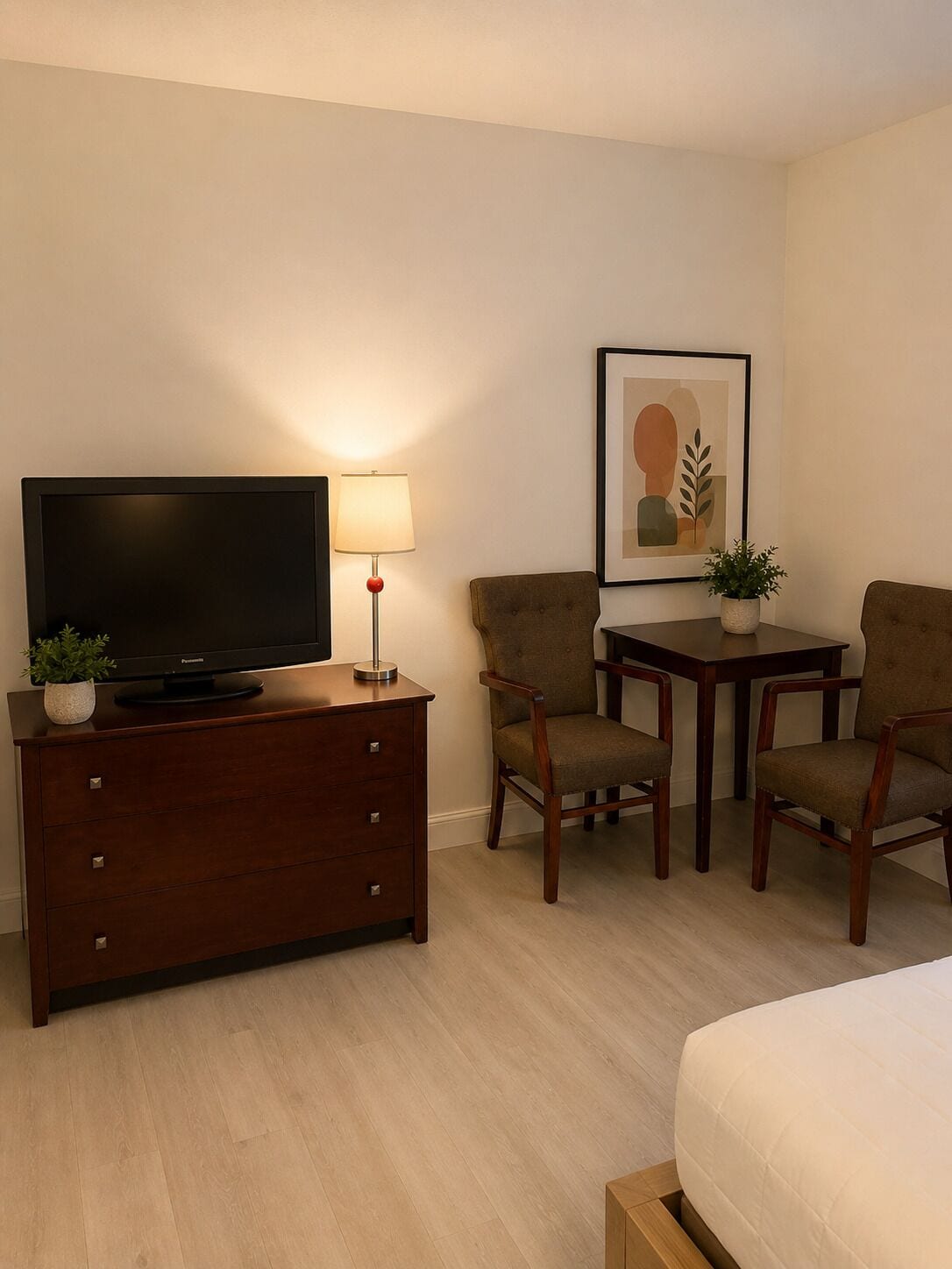 Habitación de hotel con televisor de pantalla plana sobre una cómoda de madera, dos sillas, una mesita, una obra de arte enmarcada en la pared y una cama.