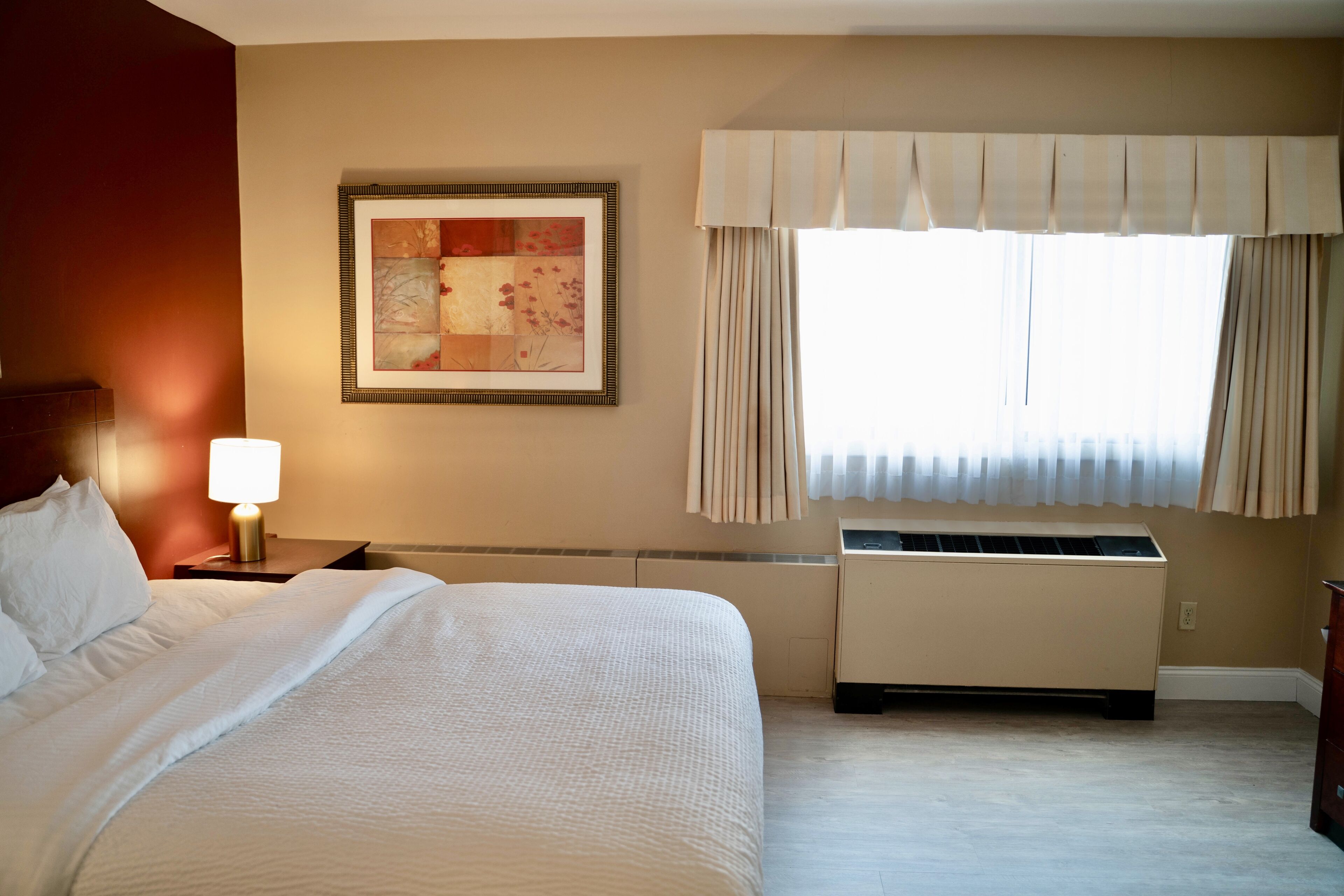 A hotel room with a bed, a nightstand with a lamp, a radiator, and a window with curtains.
