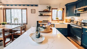 Een moderne keuken met donkerblauwe kastjes, een wit aanrecht en een marmeren eiland. Er staat een eettafel met stoelen en een bank, en een raam met uitzicht op bomen.