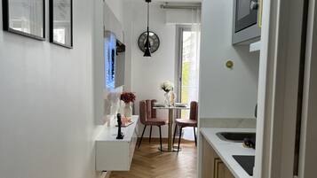 Een moderne keuken met een magnetron, spoelbak en houten kastjes. Er is een eethoek met een tafel en stoelen, en een gang met een deur naar een andere kamer.