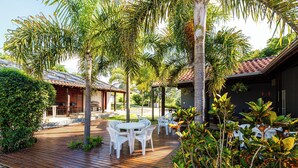 Une terrasse en bois, agrémentée de chaises et de tables blanches, entourée de palmiers et de verdure.