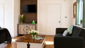A living room with a flat-screen TV mounted on a wooden panel wall, a white door, a dark sofa with cushions, and a coffee table with a vase of flowers. - Lodge 1 - Lytton Tree Lodge - Reydon (Reydon)