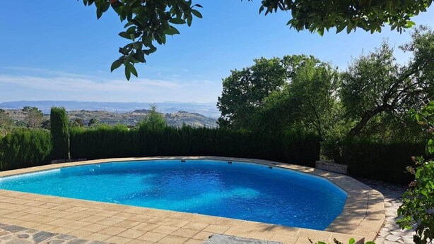Una piscina con borde de losetas de piedra, rodeada de follaje y árboles, con vistas a colinas distantes.