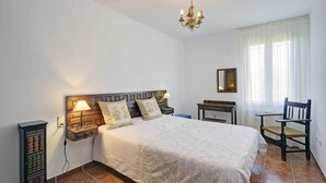 Una habitación de hotel con una cama grande, una mesita de noche con lámpara, una silla de madera, una pequeña mesa y una estantería.