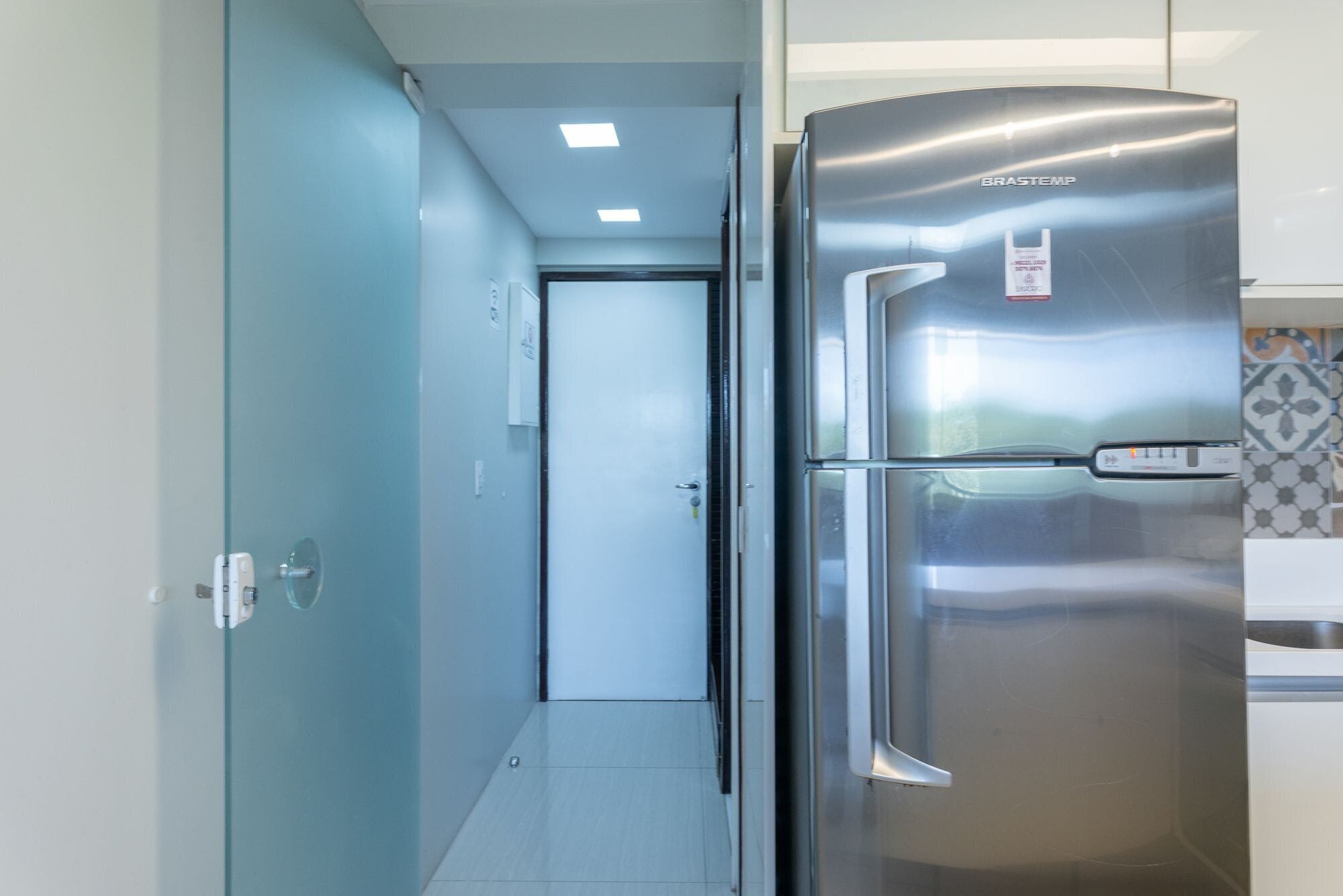Un réfrigérateur en acier inoxydable dans une cuisine avec un mur recouvert de carreaux et une porte donnant sur un couloir.