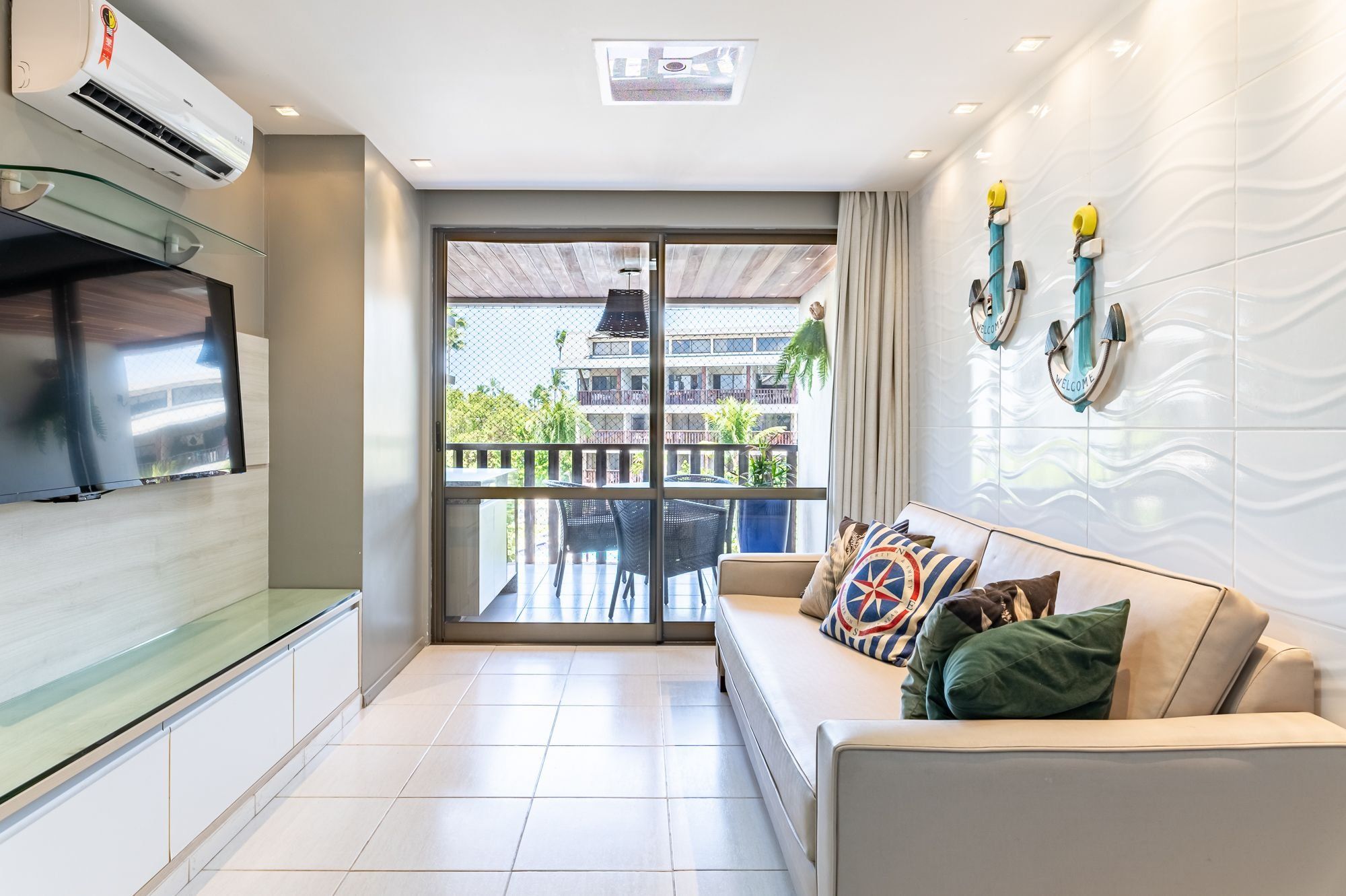 Una sala moderna con televisor de pantalla plana, un sofá con cojines estampados y un balcón con vista a un edificio y vegetación.