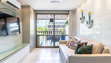 Una sala moderna con televisor de pantalla plana, un sofá con cojines estampados y un balcón con vista a un edificio y vegetación.