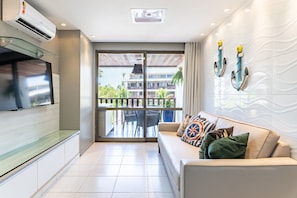 Una sala moderna con televisor de pantalla plana, un sofá con cojines estampados y un balcón con vista a un edificio y vegetación.