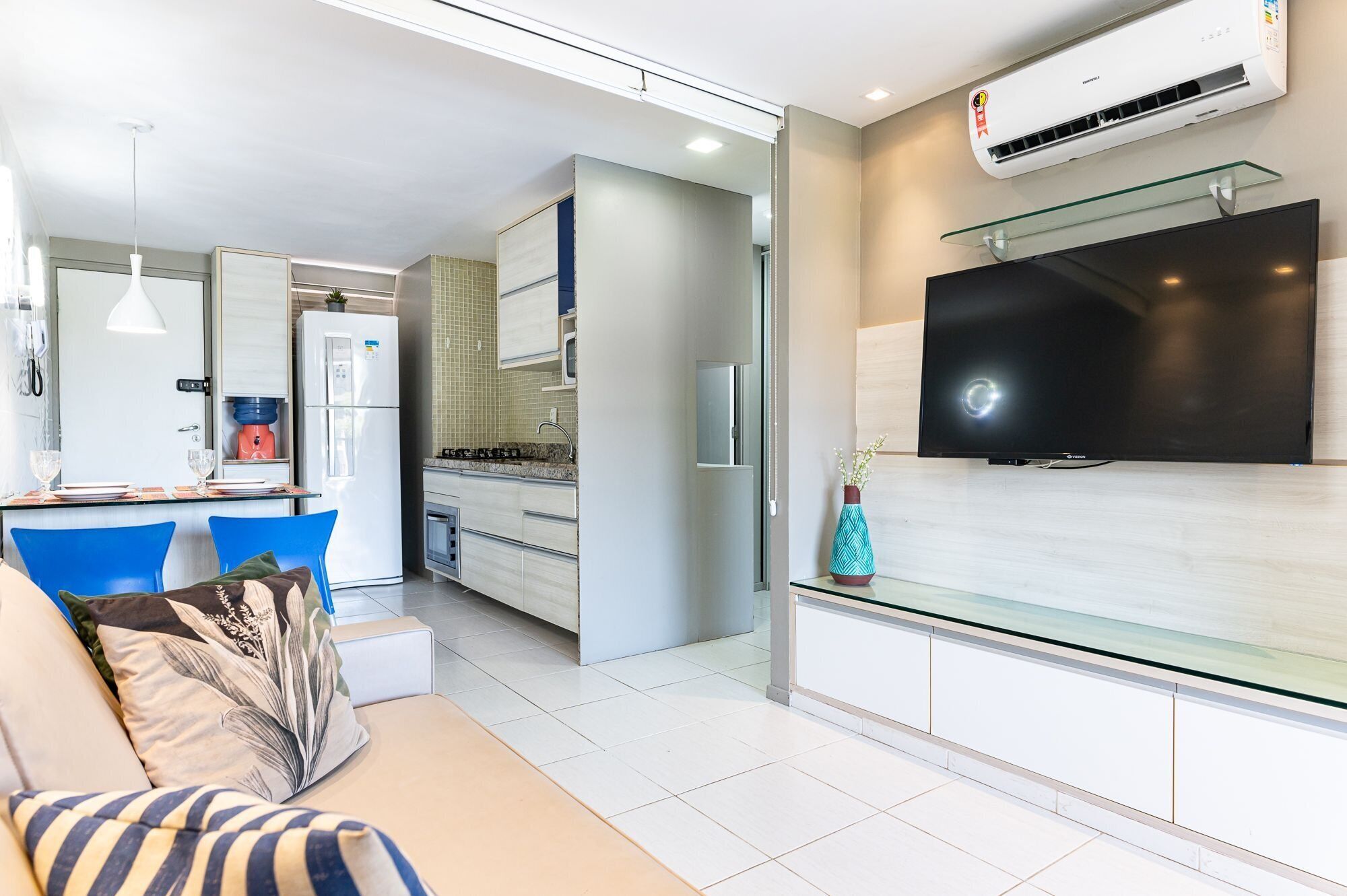 Uma sala de estar moderna com um sofá, uma televisão de tela plana fixada em uma prateleira de vidro e uma área de cozinha com geladeira e micro-ondas.