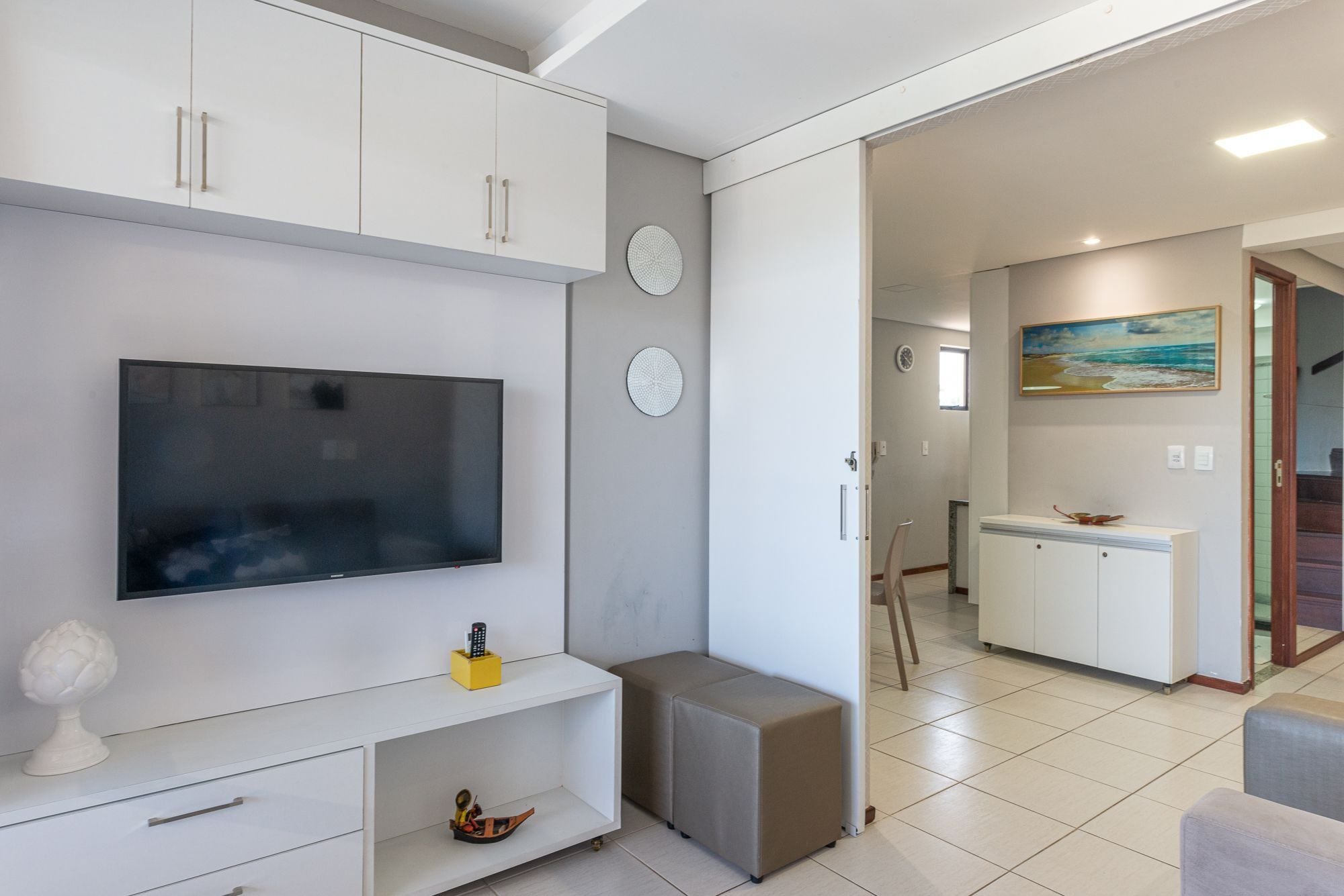 Una sala moderna con un televisor de pantalla plana, un mueble blanco, un taburete pequeño y un cuadro en la pared.