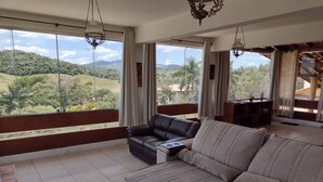 Amplia sala de estar con un ventanal que ofrece una vista panorámica de colinas y vegetación. Hay un sofá color crema y un sillón de cuero negro.