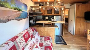 Un espacio habitable compacto con kitchenette, un sofá con cojines estampados y un mural de una playa.