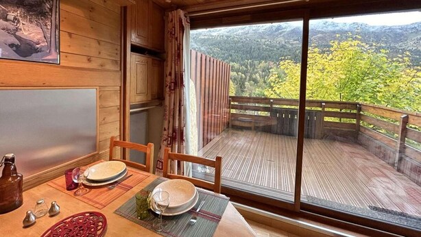 Interior de madera con una mesa para dos, una ventana grande con vistas a una cadena montañosa boscosa y un balcón con barandilla de madera.