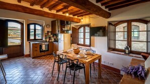 A spacious kitchen with a dining area, wooden beams, large windows, and a rustic stone wall.