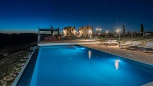 A swimming pool with lounge chairs, a covered seating area, and stone buildings in the background.