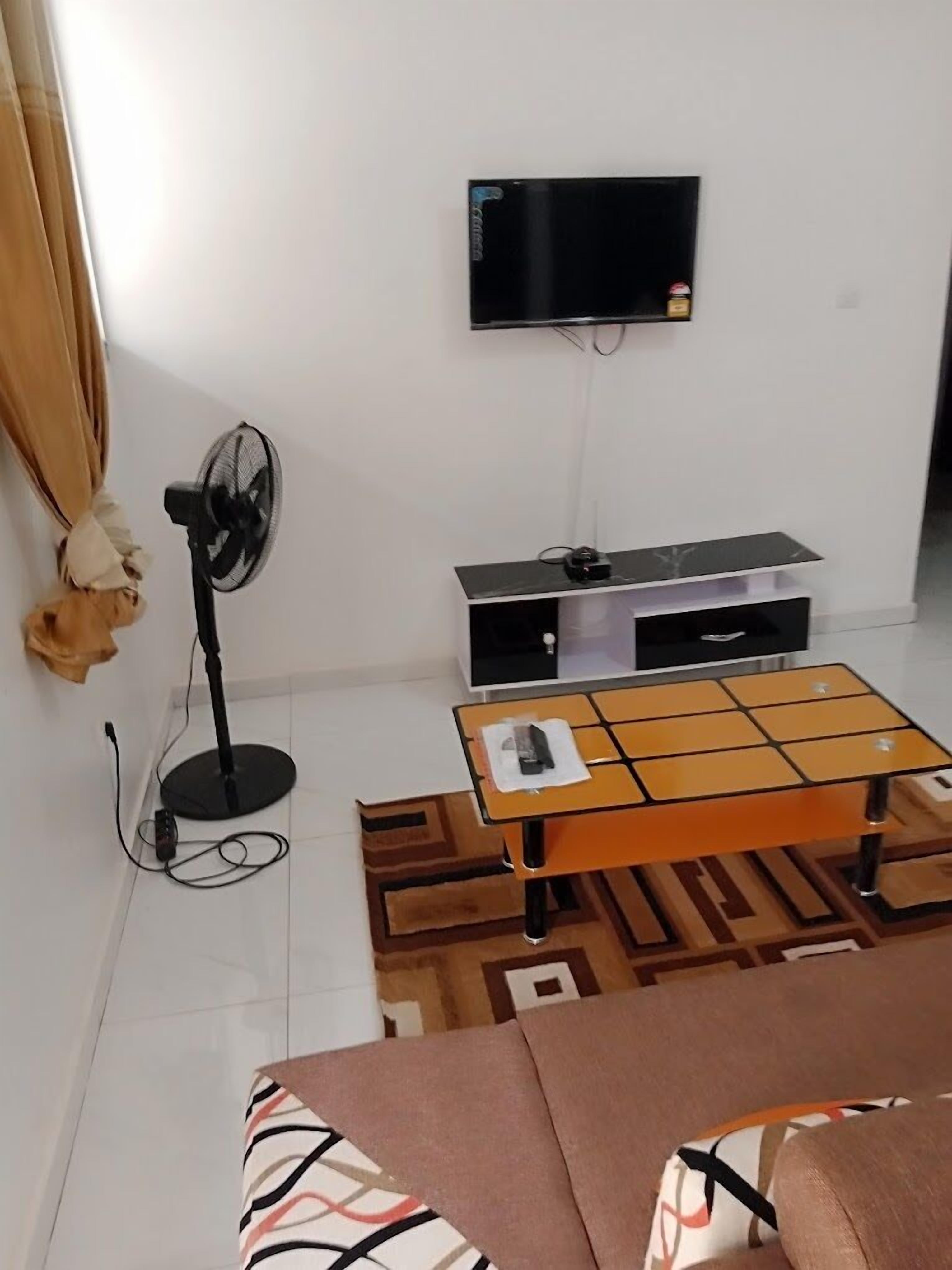 A living room with a flat-screen TV mounted on the wall, a standing fan, a wooden coffee table with a remote, and a patterned rug.