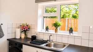 A kitchen with a stainless steel sink, a dish rack, a washing machine, and a window with a view of a wooden fence and greenery.
