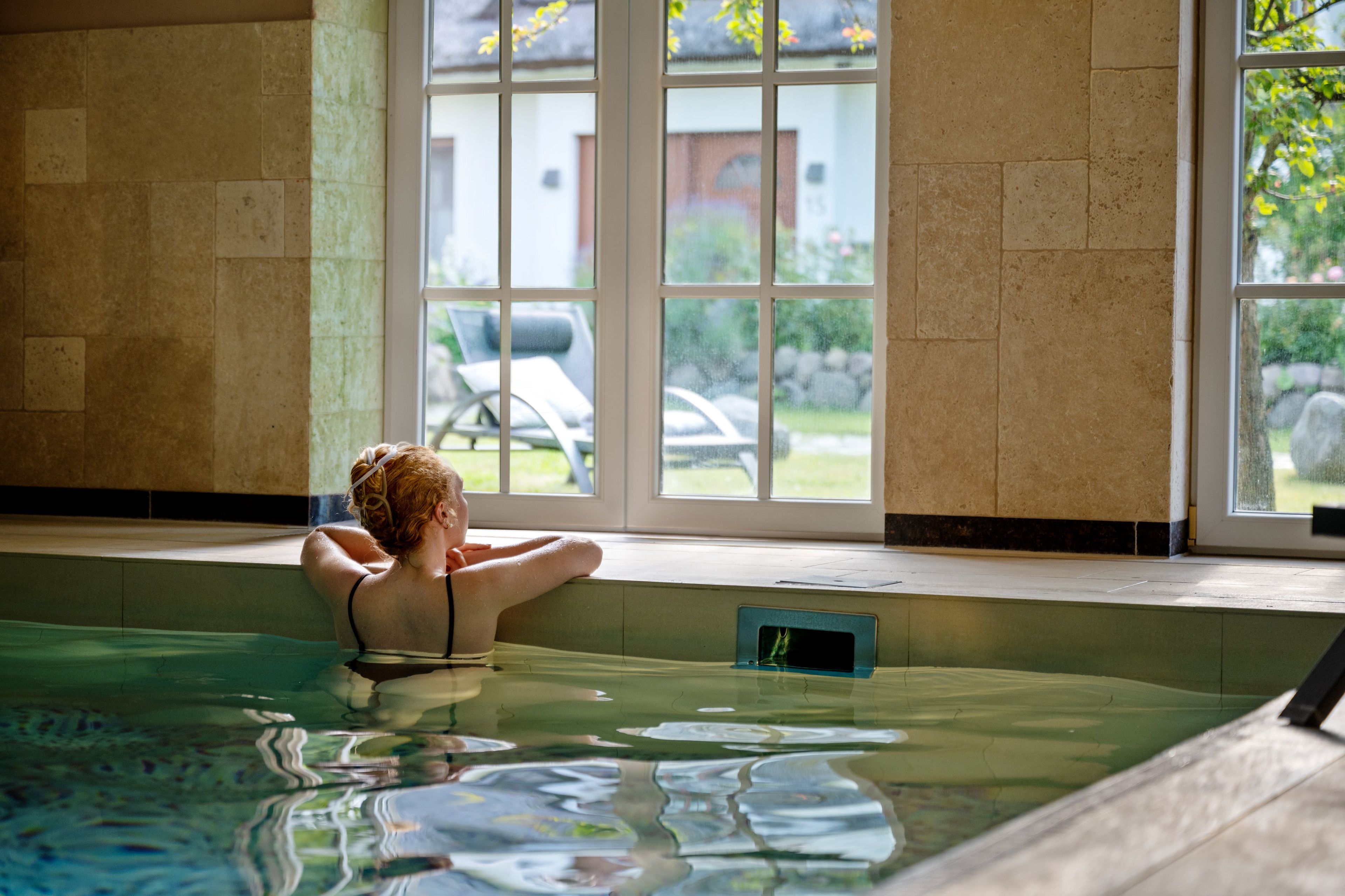 Une personne est assise près d’une fenêtre donnant sur la piscine, avec vue sur une maison et une voiture à l’extérieur.