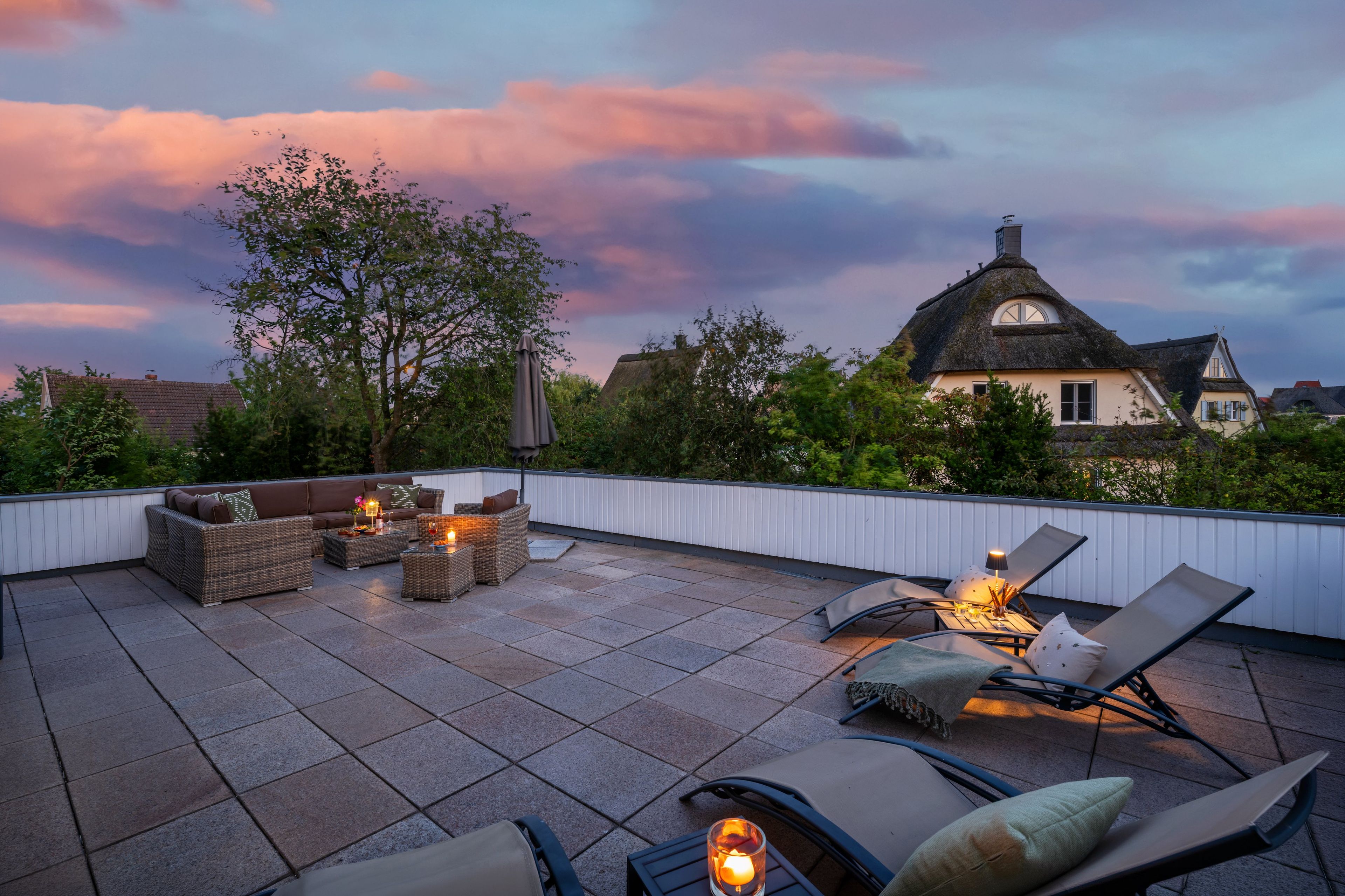 Une terrasse sur le toit, aménagée avec du mobilier d’extérieur, une table décorée de bougies et une vue sur une maison à toit de chaume.