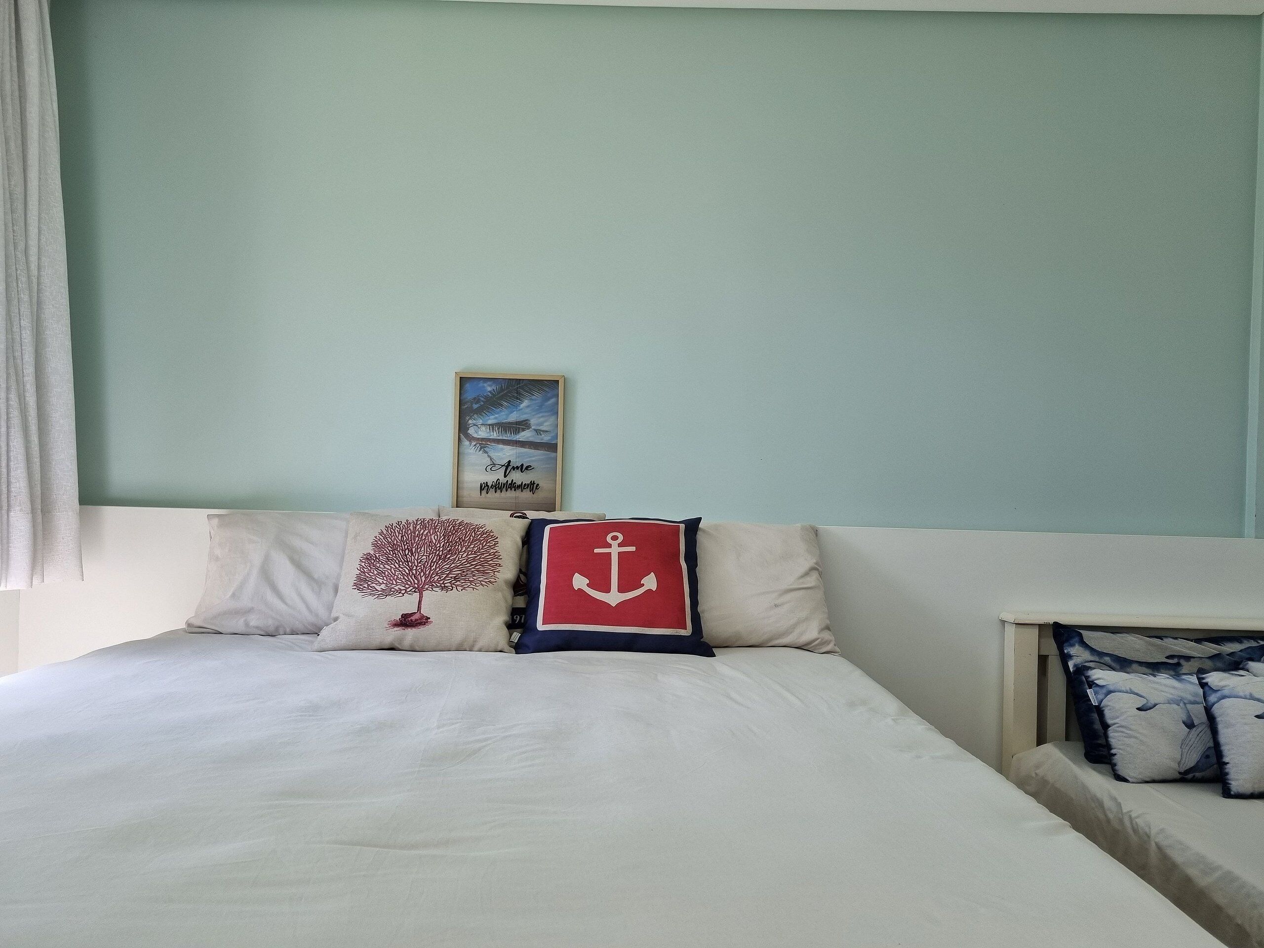 Uma cama com colchão e almofadas brancos, um quadro pendurado na parede e uma janela com cortinas.