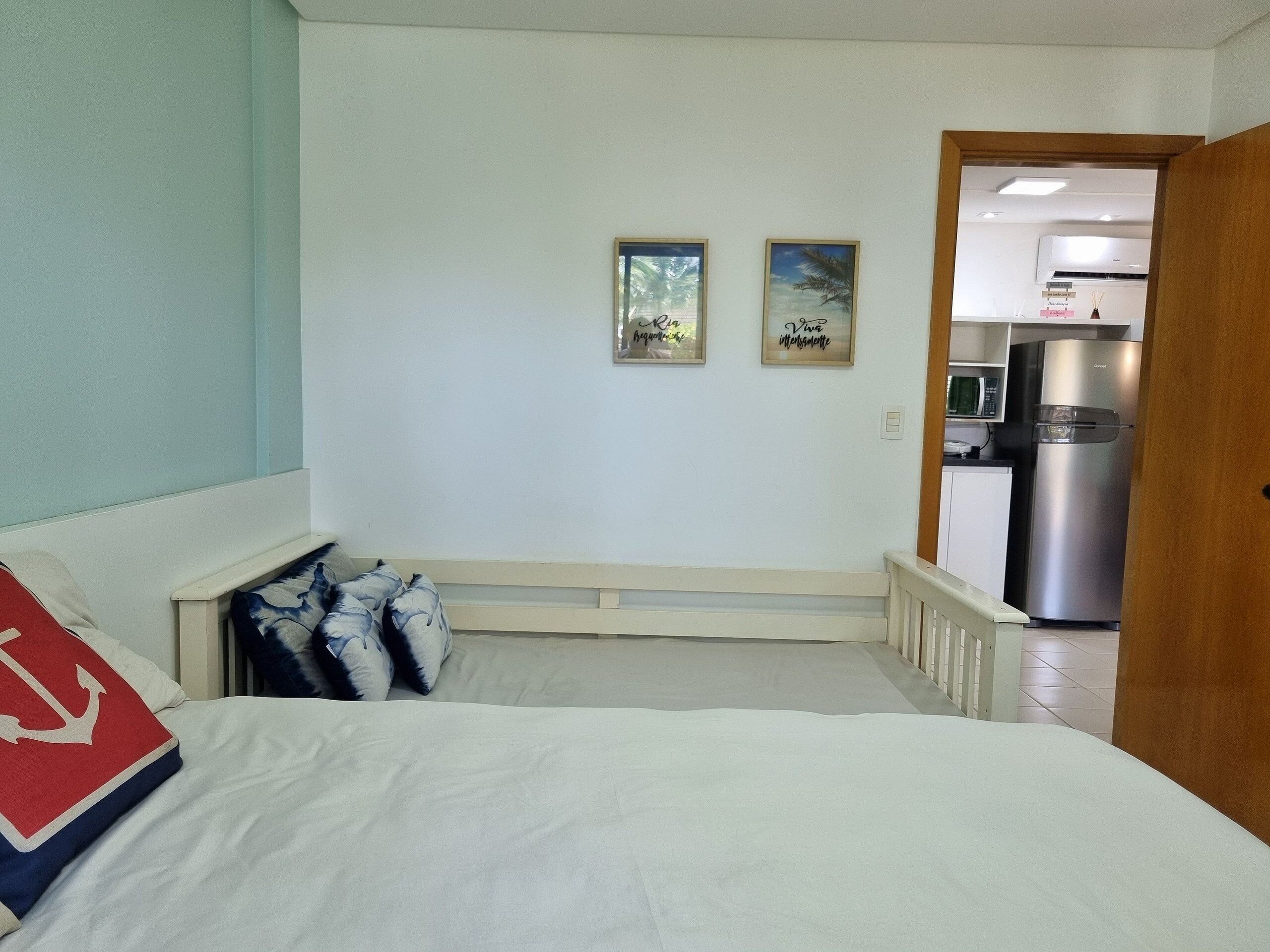 A bedroom with a bed, a nightstand, a door leading to a kitchenette, and two framed pictures on the wall.