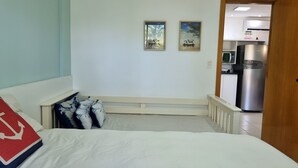 Ein Schlafzimmer mit einem Bett, einem Nachttisch, einer Tür zu einer Küchenzeile und zwei gerahmten Bildern an der Wand.