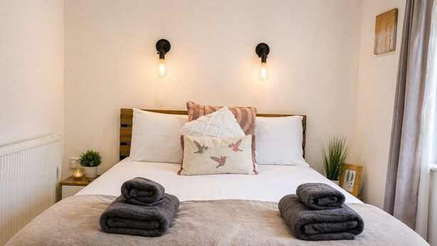 A neatly made bed with two rolled towels, a pillow with a bird design, and two wall-mounted light fixtures. - Lodge 3 - Lytton Tree Lodge (Reydon)