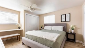 A bedroom with a bed, a bench, a bedside table, a ceiling fan, and a window with blinds. - The Sunshine House (Sequim)