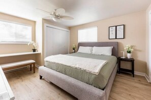 Ein Schlafzimmer mit einem Bett, einer Bank, einem Nachttisch, einem Deckenventilator und einem Fenster mit Jalousien.