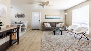 A spacious living room with a sofa, coffee table, and two white chairs. A dining area with a table and chairs is visible in the background. - The Sunshine House (Sequim)