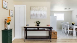 A room with a wooden floor, a dark wooden table, a white door, a white wall with a painting, and a dining area with white chairs and a table. - The Sunshine House (Sequim)