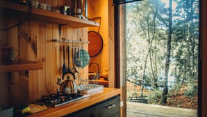 A wooden kitchen with a stove, hanging utensils, and a view of a forest through a large window.