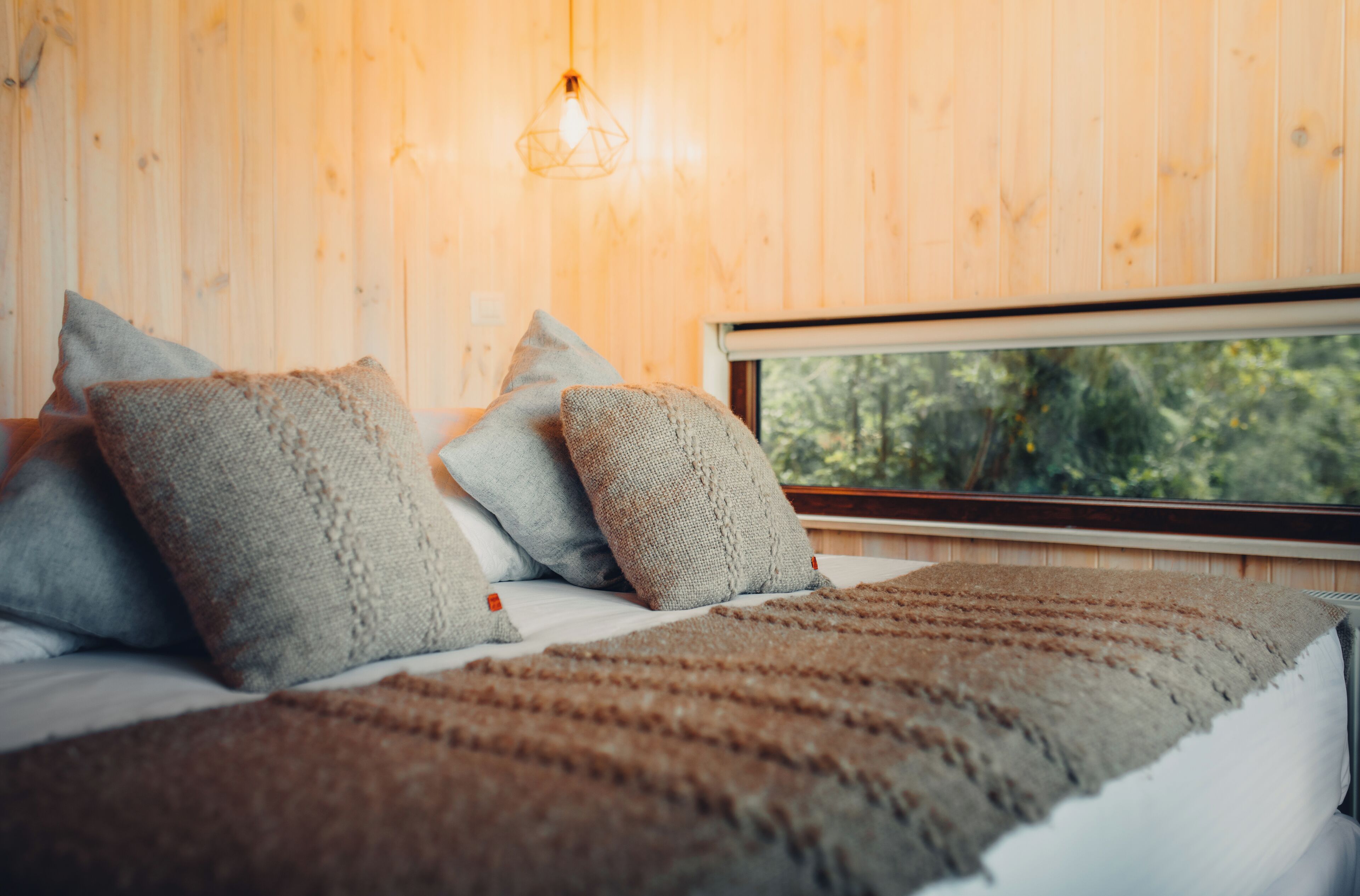 A bed with a brown blanket and grey pillows in a room with wooden walls and a window overlooking greenery.