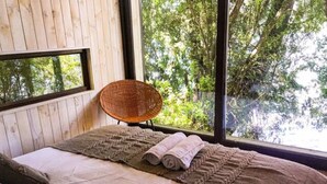 A bedroom with a large window, a woven chair, a bed with a brown blanket, and a view of trees.