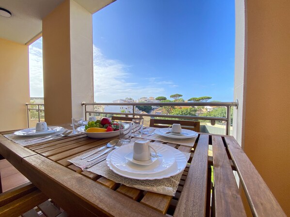 A wooden table set for two with plates, cutlery, and a bowl of fruit on a balcony with a clear view of buildings and trees.