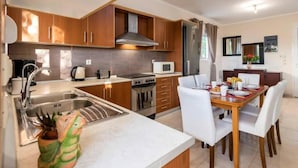 A modern kitchen with wooden cabinets, a stainless steel sink, a stove, and a dining area with white chairs and a table. - Angels Villa in Kremasti Near the Beach (Rhodes)