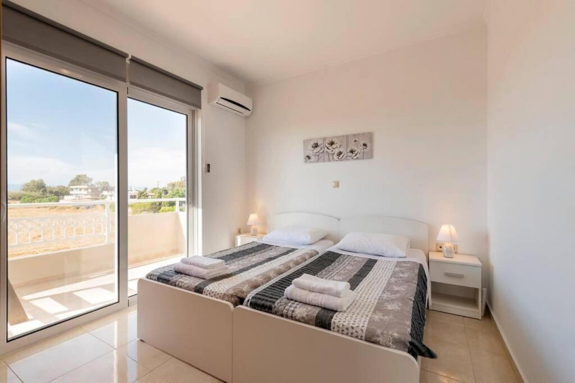 A bedroom with a large bed, two bedside tables with lamps, a wall-mounted air conditioner, and a view of the outdoors through a sliding glass door.