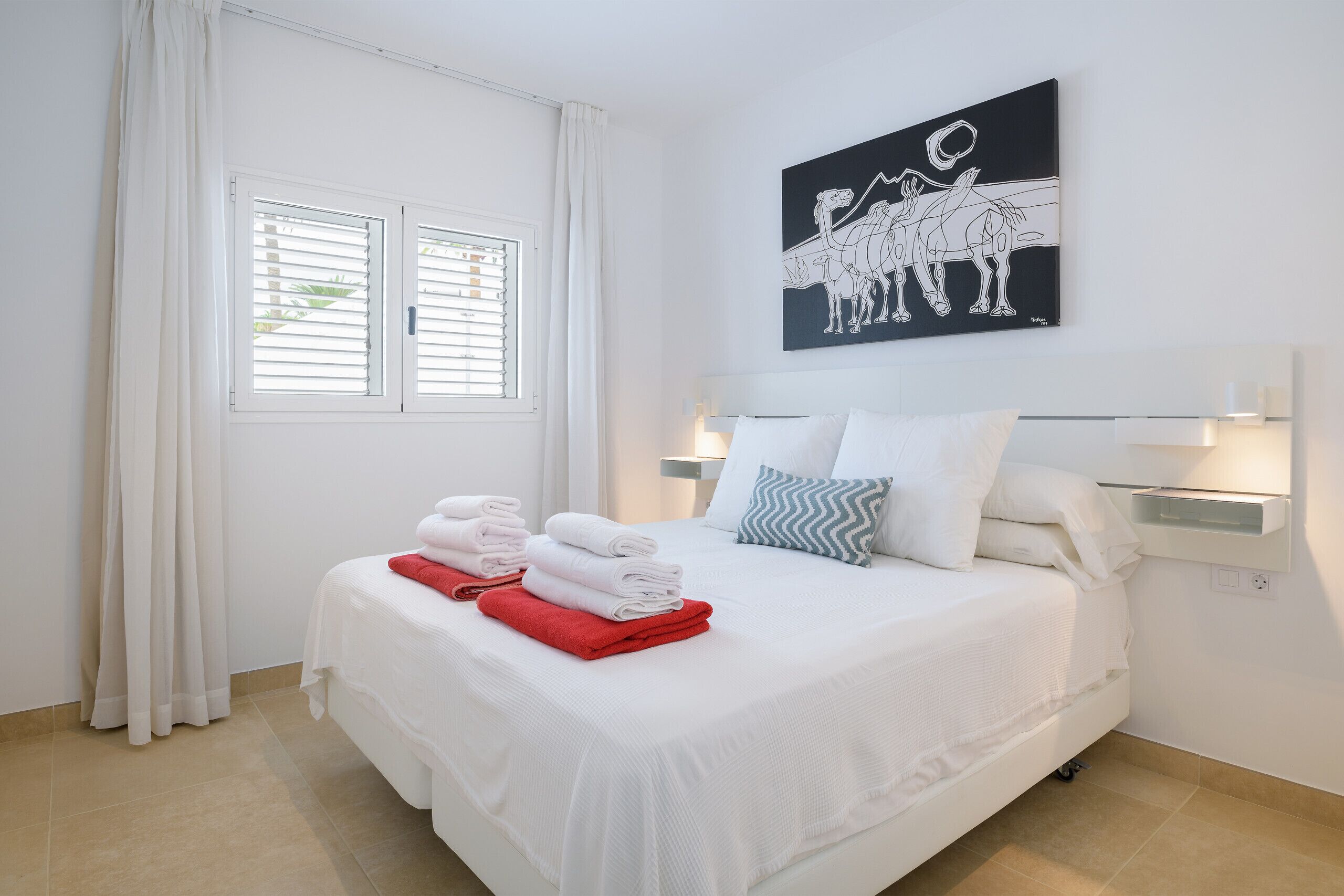 A neatly made bed with white and red towels, a black and white artwork above it, and a window with white curtains.