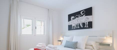 A neatly made bed with white and red towels, a black and white artwork above it, and a window with white curtains.